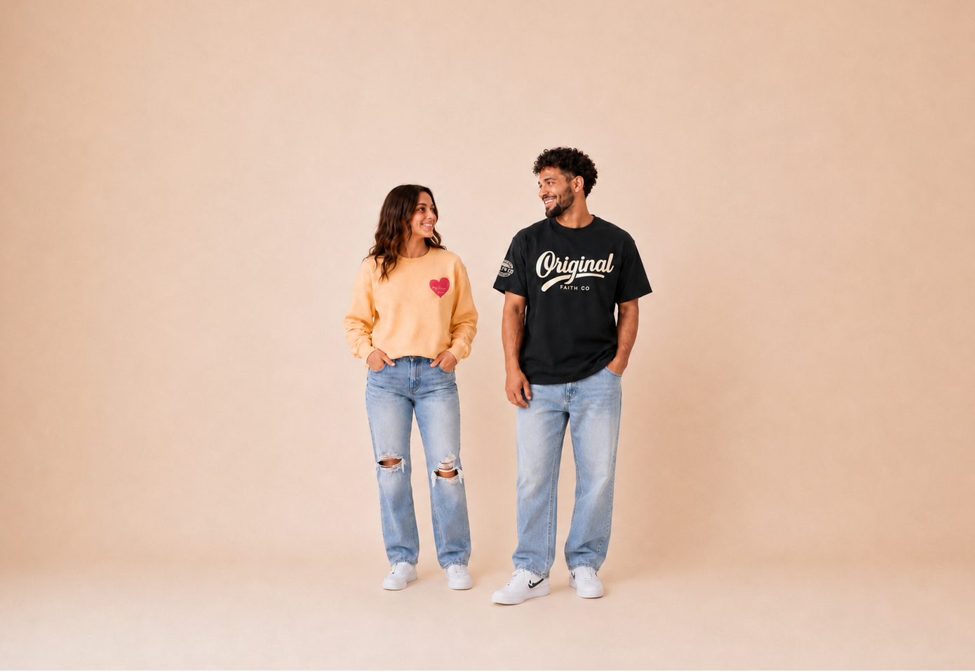 A man and a woman standing side-by-side in a studio setting, wearing matching black graphic t-shirts with distressed light-wash jeans and white sneakers against a solid beige backdrop.