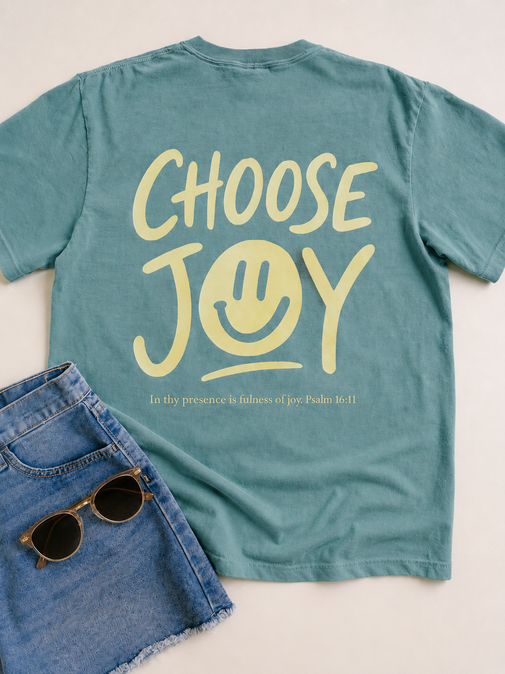 A teal green t-shirt with a yellow graphic reading 'CHOOSE JOY' and a smiley face, styled flat-lay with denim shorts and sunglasses on a light beige background.