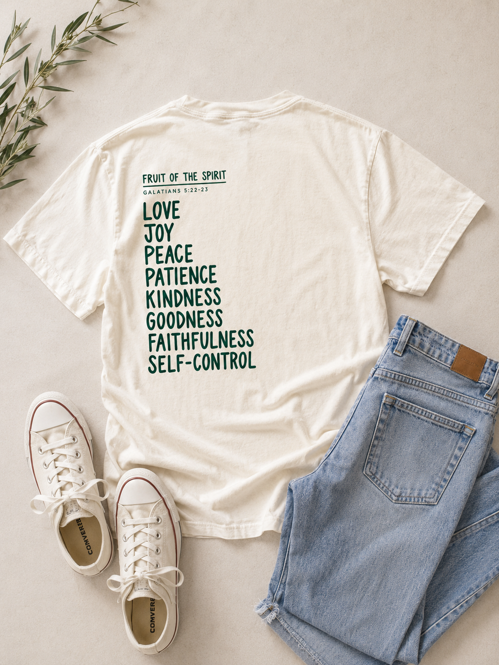 An off-white graphic t-shirt with a list of biblical virtues printed in dark green, styled in a flat lay with light blue jeans, white sneakers, and a sprig of greenery on a light beige background.