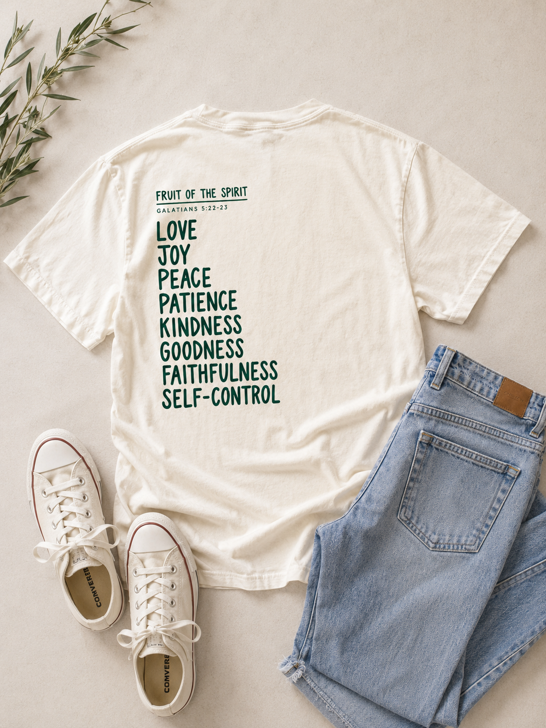 An off-white graphic t-shirt with a list of biblical virtues printed in dark green, styled in a flat lay with light blue jeans, white sneakers, and a sprig of greenery on a light beige background.
