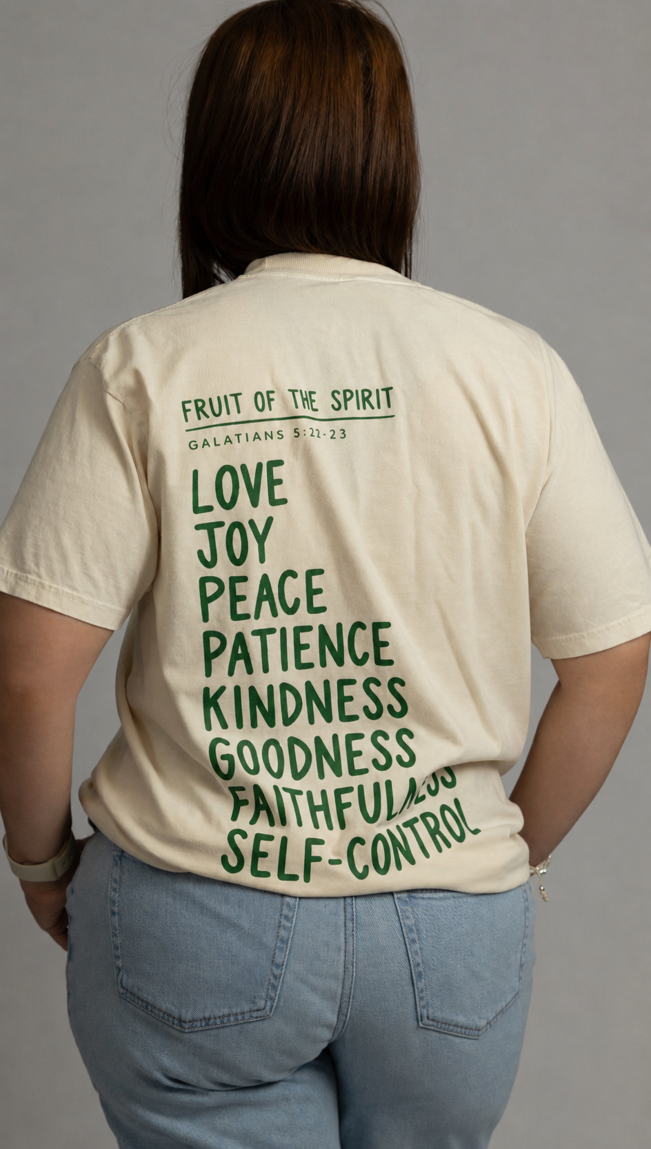 A person shown from the back wearing a cream-colored t-shirt with green text listing virtues like love, joy, peace, patience, kindness, goodness, faithfulness, and self-control, paired with light blue jeans against a plain gray background.