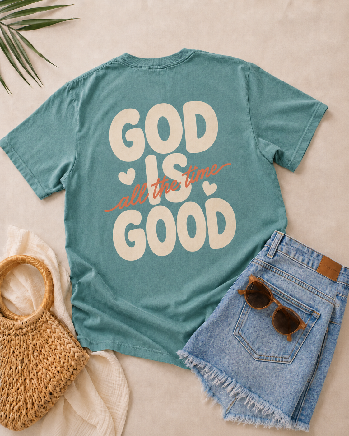 A styled flat-lay of a teal short-sleeve T-shirt with the phrase 'GOD IS all the time GOOD' printed on the back in cream and orange. The shirt is arranged on a light beige fabric with denim shorts, sunglasses, a woven bag, and a leaf in the corner.